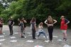 Mehrere Jugendliche stehen auf einem Kiesplatz im Freien, verteilt auf einzelne Zeitungen, umgeben von Bäumen